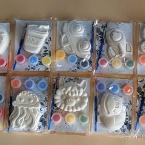 Op de afbeelding: Tien individuele knutselpakketten in doorzichtige plastic verpakking, elk met een wit gipsfiguur, een klein penseel en drie kleine verfpotjes. De figuren omvatten een donut, een colabeker, een auto, een raket, een schildpad, een fiets, een kwal, een leeuw, een nijlpaard en patat.