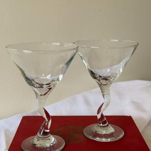 May include: Two clear martini glasses with a unique twisted stem design. The stems feature a red accent, and the glasses sit on a red book. The glasses have a classic, elegant shape, perfect for cocktails.