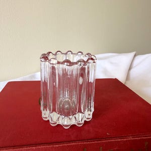 May include: A clear, cylindrical glass votive candle holder with vertical ribbed detailing. The top edge features a scalloped design. The candle holder is displayed on a red book.