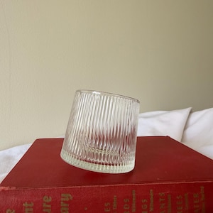 May include: A clear, angled glass tumbler with vertical ribbed detailing. The glass sits on a vintage red book titled "The Giant Nature Library". The book's spine features gold lettering and titles.