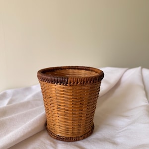 May include: A small, cylindrical woven basket with a natural brown color. The basket has a darker brown trim around the top and base. The weave is tight and even, creating a sturdy container. The basket is likely used for storage or decoration.
