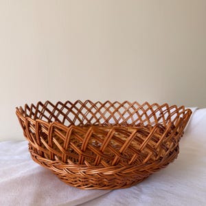 May include: A round, woven basket with a lattice design. The basket is made of brown wicker and sits on a white cloth. The basket's rim has a scalloped edge.