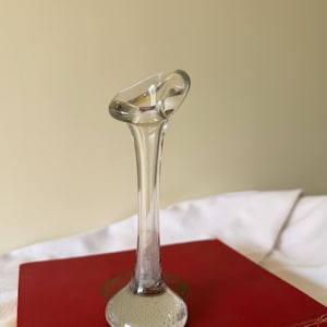 May include: A clear glass bud vase with a flared top and a bulbous base. The vase is set on a red book. The vase has a long, slender neck and a slightly tilted opening. The base is filled with tiny bubbles.