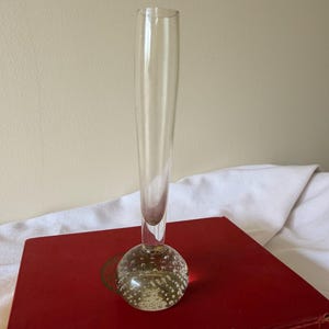 May include: A clear glass bud vase with a round, bubble-textured base and a slender, cylindrical neck. The vase is displayed on a red book, showcasing its elegant design. The vase is ideal for displaying a single flower or small floral arrangement.