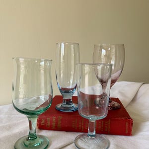 May include: Four clear glass goblets with colored stems and bases. The goblets are different shapes and sizes, with blue and green stems. The goblets are arranged on top of a red book.