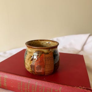 May include: A small, handcrafted ceramic mug with a textured, earthy design. The mug features patches of brown, orange, and green. It sits atop a vintage red book with gold lettering. The mug is approximately 3 inches in diameter and 2.5 inches tall.