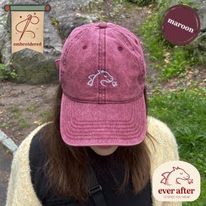 Può includere: Cappellino da baseball bordeaux con un design a cavallo ricamato bianco. Il cappellino ha una visiera curva e un cinturino regolabile. La parola "embroidered" è stampata su un'etichetta. Il logo del marchio è visibile in basso a destra.