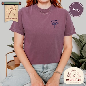 Könnte beinhalten: Beerenfarbenes T-Shirt mit einem kleinen, gestickten Augendesign. Das Shirt hat kurze Ärmel und einen Rundhalsausschnitt. Das Wort "embroidered" befindet sich auf einem Banner oben links. Das Markenlogo befindet sich unten rechts.
