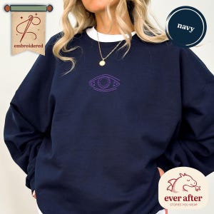 Può includere: Felpa blu navy con colletto bianco e un design a occhio ricamato viola. La felpa ha la parola "embroidered" su uno striscione in alto a sinistra e le parole "ever after stories you wear" su un logo in basso a destra.