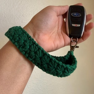 May include: A dark green, crocheted wristlet key holder. The wristlet is made of a thick, textured yarn and is attached to a metal clasp. A black car key fob is attached to the clasp.
