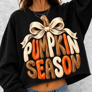 Pode incluir: Uma camisola preta de manga comprida e corte curto com um gráfico de uma abóbora rematada por um grande laço bege. O texto PUMPKIN SEASON está impresso por baixo numa fonte arredondada e estilizada em tons de laranja, castanho, creme e branco.