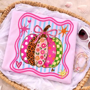 May include: A light pink t-shirt featuring a central pumpkin graphic with multicolored segments including leopard print, floral patterns, polka dots, and stars. The pumpkin is topped with a pink bow and surrounded by a wavy pink border and blue stripes.