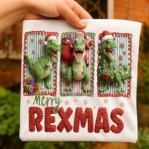 May include: White t-shirt with a festive design featuring three green crocheted dinosaurs wearing Santa hats and Christmas lights. The text "Merry REXMAS" is displayed in red, festive lettering. The design includes presents and snowflakes.