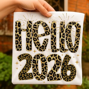 Puede incluir: Camiseta blanca con la frase "HELLO 2026" en un diseño de estampado de leopardo dorado y negro. El diseño incluye detalles de purpurina dorada y estrellas, creando un aspecto festivo.