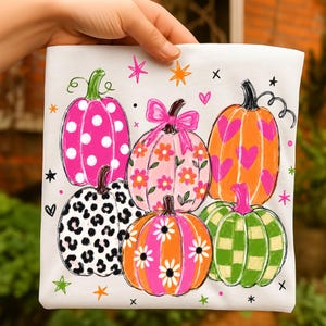 May include: A white t-shirt featuring a colorful graphic print of five pumpkins. The pumpkins display various patterns including white polka dots on hot pink, pink flowers on light pink, pink hearts on orange, black leopard print, and green stripes.