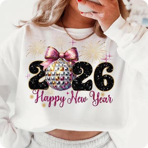 Puede incluir: Sudadera blanca con un diseño festivo para el Año Nuevo. El gráfico presenta los números "2026" en negro, una bola de discoteca, un lazo rosa y las palabras "Happy New Year" en escritura rosa, con fuegos artificiales dorados.