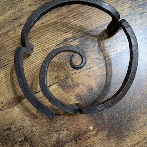 May include: A dark, wrought iron decorative piece with a spiral design. The metalwork features a central spiral and a surrounding circular shape, connected by curved segments. The piece has a rustic, handmade appearance.