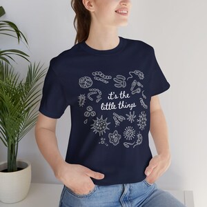 Op de afbeelding: Marineblauwe t-shirt met een witte afbeelding van verschillende micro-organismen in een cirkelvormig patroon. De tekst "it's the little things" is eronder in het wit gedrukt. Het shirt is gemaakt van een zacht materiaal.