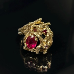 May include: A gold ring featuring a textured, branch-like metal band with an organic, gnarled appearance. The ring is set with a prominent oval-cut deep red ruby and two smaller, round-cut red rubies embedded within the intricate metal structure.