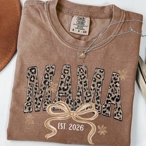 Könnte beinhalten: Ein braunes Comfort Colors T-Shirt mit dem Wort "MAMA" in Leopardenmuster-Buchstaben. Unter dem Wort "MAMA" befindet sich eine Schleife und der Text "EST. 2026". Eine silberne Halskette mit Anhänger liegt über dem Shirt.