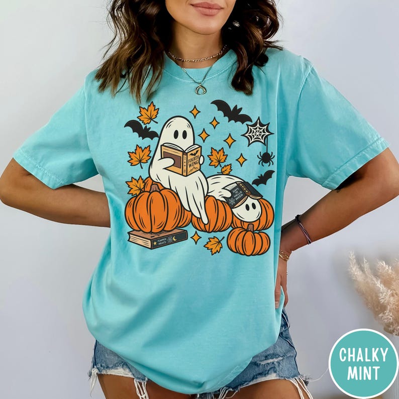 K&ouml;nnte beinhalten: Ein hellblaues T-Shirt mit einer Grafik von zwei wei&szlig;en Geistern, die B&uuml;cher lesen und auf orangefarbenen K&uuml;rbissen sitzen. Das Design enth&auml;lt orangefarbene Herbstbl&auml;tter, schwarze Flederm&auml;use, ein Spinnennetz und eine Spinne. Das Buch tr&auml;gt den Titel Fourth Wing.