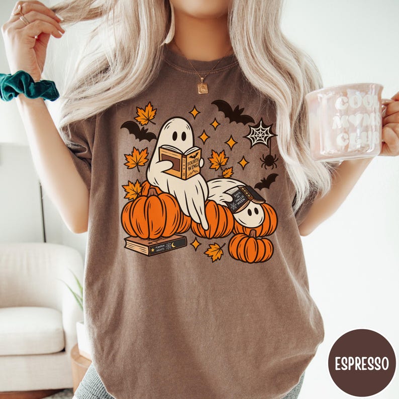 K&ouml;nnte beinhalten: Ein espressofarbenes T-Shirt mit einem Grafikdruck von zwei wei&szlig;en Geistern, die B&uuml;cher lesen und zwischen orangefarbenen K&uuml;rbissen sitzen. Das Design enth&auml;lt gelbe Ahornbl&auml;tter, schwarze Flederm&auml;use, ein Spinnennetz und eine Spinne.