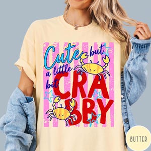May include: A pale yellow t-shirt featuring a graphic print with pink vertical stripes, two yellow crabs, and the text "Cute but a little bit CRABBY" in blue and red lettering, accented with small blue stars throughout the design.