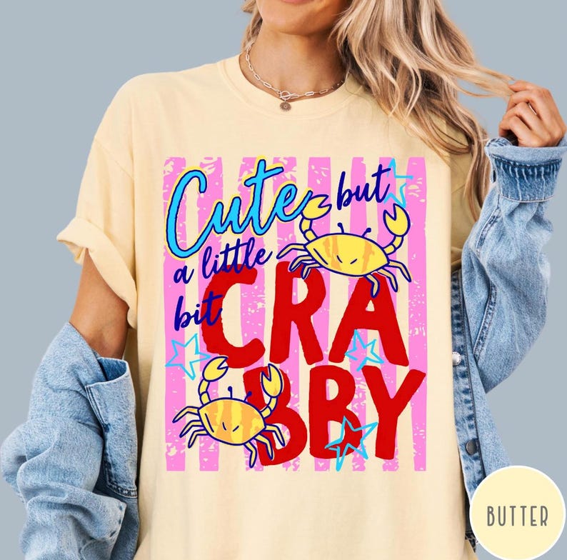May include: A pale yellow t-shirt featuring a graphic print with pink vertical stripes, two yellow crabs, and the text "Cute but a little bit CRABBY" in blue and red lettering, accented with small blue stars throughout the design.