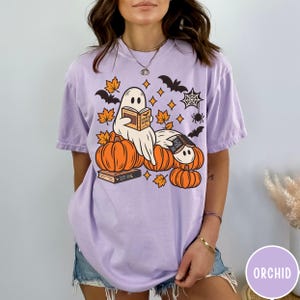 K&ouml;nnte beinhalten: Ein hellviolettes Kurzarm-T-Shirt mit einer Grafik von zwei Geistern, die B&uuml;cher lesen und auf orangefarbenen K&uuml;rbissen sitzen. Das Design enth&auml;lt orangefarbene Ahornbl&auml;tter, schwarze Flederm&auml;use, ein Spinnennetz und eine Spinne.