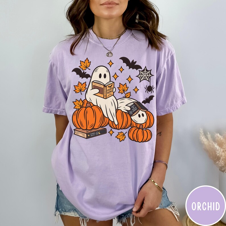 K&ouml;nnte beinhalten: Ein hellviolettes Kurzarm-T-Shirt mit einer Grafik von zwei Geistern, die B&uuml;cher lesen und auf orangefarbenen K&uuml;rbissen sitzen. Das Design enth&auml;lt orangefarbene Ahornbl&auml;tter, schwarze Flederm&auml;use, ein Spinnennetz und eine Spinne.