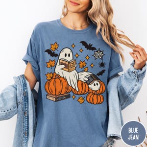 K&ouml;nnte beinhalten: Ein blaues T-Shirt mit einer Grafik von zwei wei&szlig;en Geistern, die zwischen orangefarbenen K&uuml;rbissen B&uuml;cher lesen. Das Design enth&auml;lt orangefarbene Herbstbl&auml;tter, schwarze Flederm&auml;use, ein Spinnennetz und eine Spinne. Auf den Buchcovern steht der Text Fourth Wing.