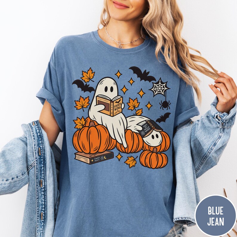 K&ouml;nnte beinhalten: Ein blaues T-Shirt mit einer Grafik von zwei wei&szlig;en Geistern, die zwischen orangefarbenen K&uuml;rbissen B&uuml;cher lesen. Das Design enth&auml;lt orangefarbene Herbstbl&auml;tter, schwarze Flederm&auml;use, ein Spinnennetz und eine Spinne. Auf den Buchcovern steht der Text Fourth Wing.