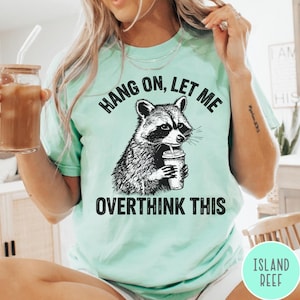 Puede incluir: Una camiseta de manga corta en color verde menta claro con una ilustración gráfica en negro de un mapache sosteniendo una taza de café. El texto sobre el mapache dice HANG ON, LET ME y el texto debajo dice OVERTHINK THIS en letras negritas.