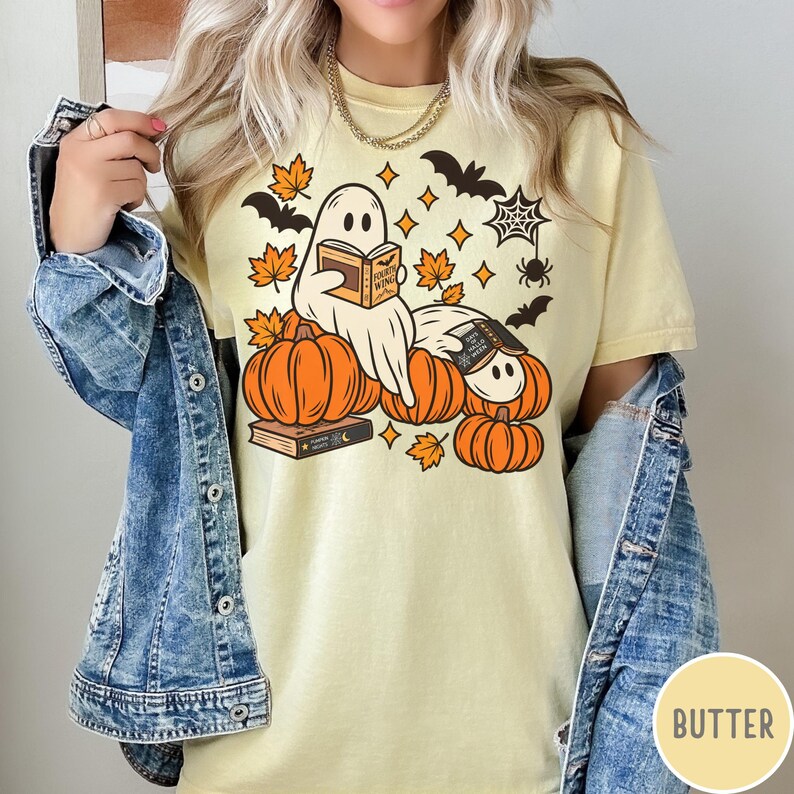 K&ouml;nnte beinhalten: Ein blassgelbes T-Shirt mit einer Grafik von zwei wei&szlig;en Geistern, die zwischen orangefarbenen K&uuml;rbissen und Herbstbl&auml;ttern B&uuml;cher lesen. Das Design enth&auml;lt schwarze Flederm&auml;use, ein Spinnennetz und kleine Sterne. Die Buchtitel lauten Fourth Wing.