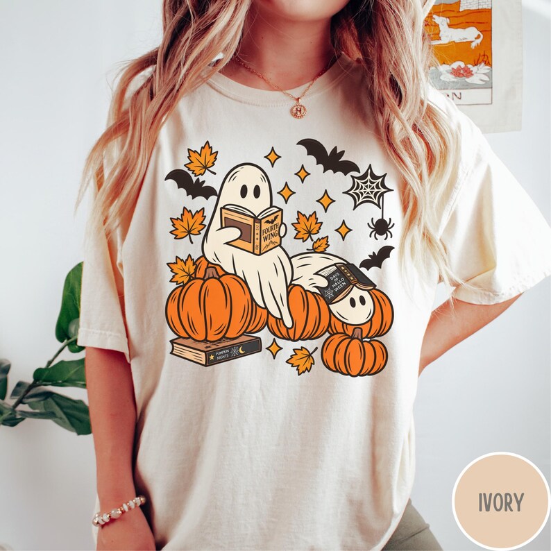 K&ouml;nnte beinhalten: Ein elfenbeinfarbenes T-Shirt mit einer Grafik von zwei Geistern, die auf orangefarbenen K&uuml;rbissen sitzen und B&uuml;cher lesen. Das Design enth&auml;lt orangefarbene Herbstbl&auml;tter, schwarze Flederm&auml;use, ein Spinnennetz und eine Spinne. Die B&uuml;cher tragen die Aufschriften Fourth Wing und Pumpkin Nights.