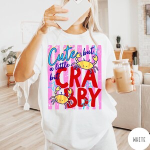 May include: A white t-shirt featuring a graphic design with pink vertical stripes, blue text reading Cute but a little bit, and large red text reading CRABBY. The design includes two yellow crab illustrations and several small blue stars.