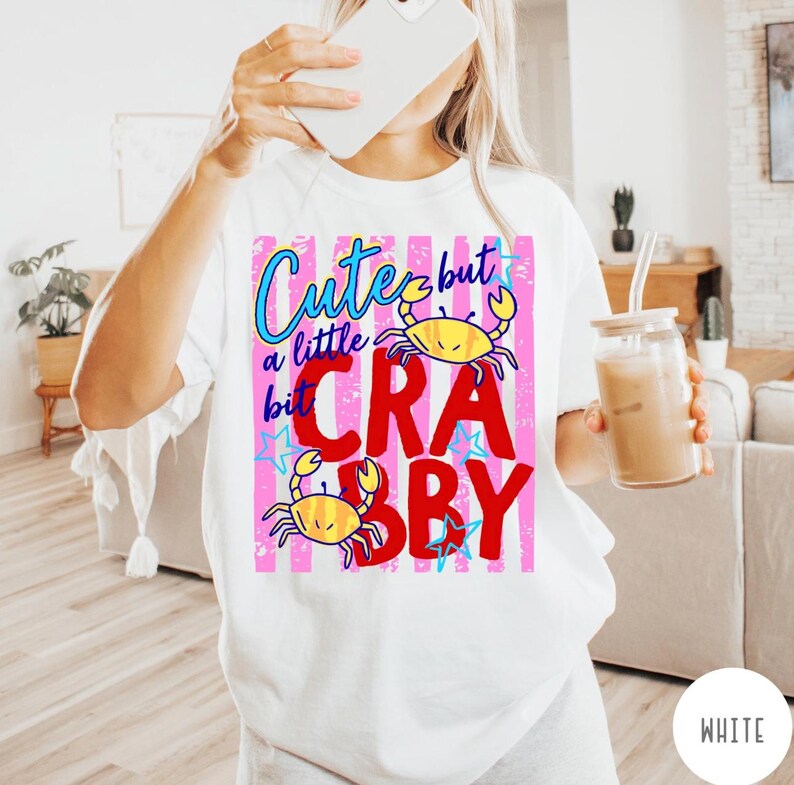 May include: A white t-shirt featuring a graphic design with pink vertical stripes, blue text reading Cute but a little bit, and large red text reading CRABBY. The design includes two yellow crab illustrations and several small blue stars.