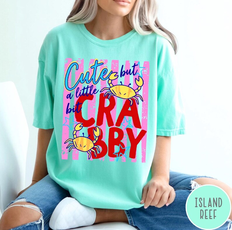 May include: A mint green short-sleeve t-shirt featuring a graphic print with the text "Cute but a little bit CRABBY" in blue and red. The design includes two yellow crabs and pink vertical stripes with blue star accents on the front.