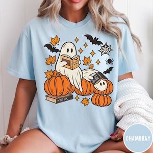 K&ouml;nnte beinhalten: Ein hellblaues T-Shirt mit einer Grafik von zwei Geistern, die zwischen orangefarbenen K&uuml;rbissen B&uuml;cher lesen. Das Design enth&auml;lt orangefarbene Ahornbl&auml;tter, schwarze Flederm&auml;use, ein Spinnennetz sowie B&uuml;cher mit der Aufschrift Fourth Wing und Days of Halloween.