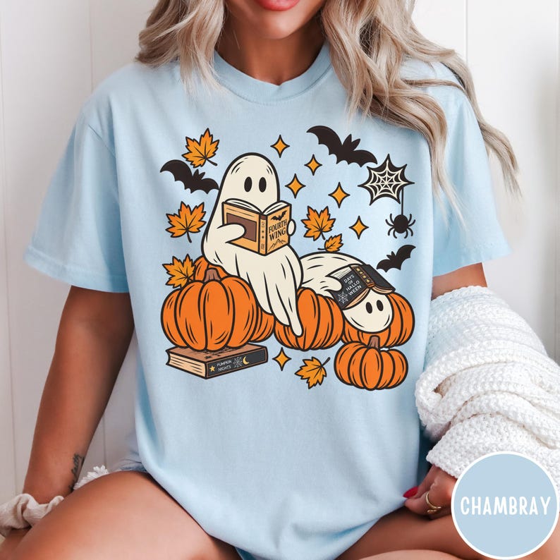 K&ouml;nnte beinhalten: Ein hellblaues T-Shirt mit einer Grafik von zwei Geistern, die zwischen orangefarbenen K&uuml;rbissen B&uuml;cher lesen. Das Design enth&auml;lt orangefarbene Ahornbl&auml;tter, schwarze Flederm&auml;use, ein Spinnennetz sowie B&uuml;cher mit der Aufschrift Fourth Wing und Days of Halloween.