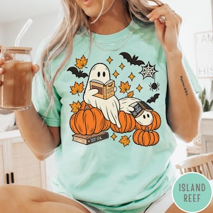 K&ouml;nnte beinhalten: Ein helles mintgr&uuml;nes T-Shirt mit einem Grafikdruck von zwei wei&szlig;en Geistern, die B&uuml;cher lesen und zwischen orangefarbenen K&uuml;rbissen sitzen. Das Design enth&auml;lt schwarze Flederm&auml;use, gelbe Sternakzente, orangefarbene Ahornbl&auml;tter und ein Spinnennetz.