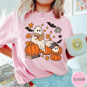 K&ouml;nnte beinhalten: Ein hellrosa T-Shirt mit einer Grafik von zwei wei&szlig;en Geistern, die B&uuml;cher lesen und auf orangefarbenen K&uuml;rbissen sitzen. Das Design enth&auml;lt orangefarbene Ahornbl&auml;tter, schwarze Flederm&auml;use, ein Spinnennetz und eine Spinne.