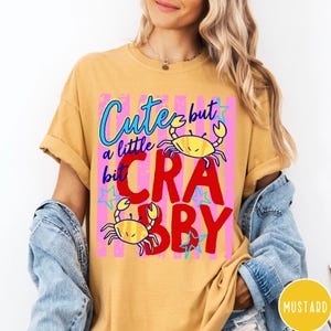 May include: A mustard yellow short-sleeve t-shirt featuring a graphic print with two yellow crabs, blue stars, and pink vertical stripes. The text reads "Cute but a little bit CRABBY" in blue and red lettering. The shirt is worn under a denim jacket.