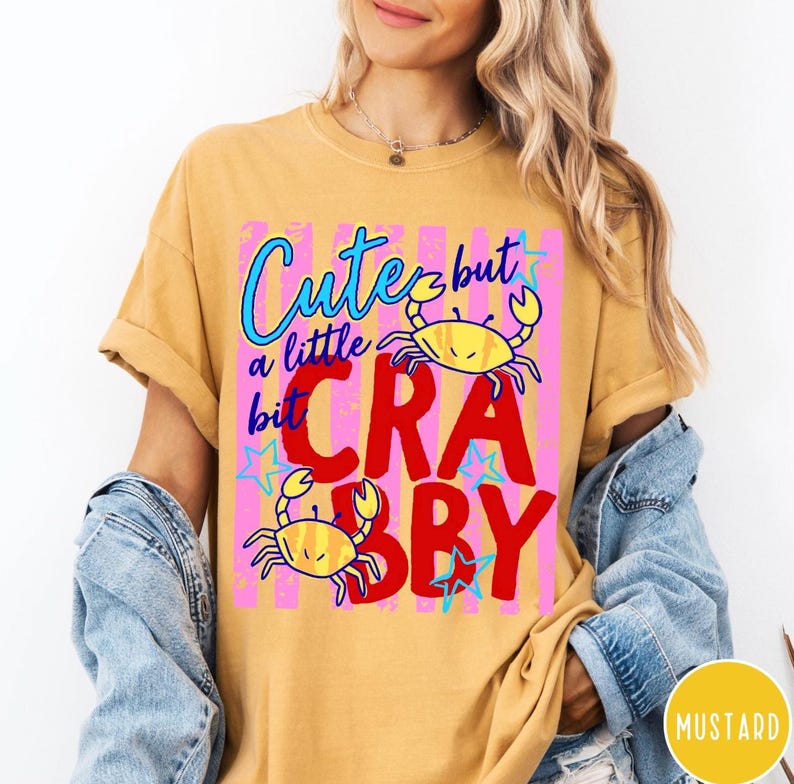 May include: A mustard yellow short-sleeve t-shirt featuring a graphic print with two yellow crabs, blue stars, and pink vertical stripes. The text reads "Cute but a little bit CRABBY" in blue and red lettering. The shirt is worn under a denim jacket.