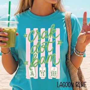 May include: A lagoon blue t-shirt featuring a graphic print with vertical pink stripes, green stars, and the text "crak dot bam" in a cursive green font. Below the text are three mahjong tiles displaying the numbers 5, 7, and 3 in green.