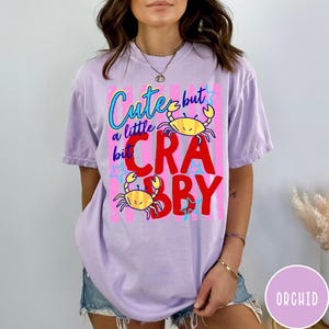 May include: A light purple short-sleeve t-shirt featuring a graphic print with the text "Cute but a little bit CRABBY" in blue and red. The design includes two yellow crab illustrations and small blue star accents on a vertical striped background.