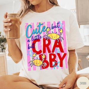 May include: An ivory t-shirt featuring a graphic print with pink vertical stripes, blue stars, and two yellow crabs. The text reads "Cute but a little bit CRABBY" in blue and red lettering. The design is centered on the front of the garment.