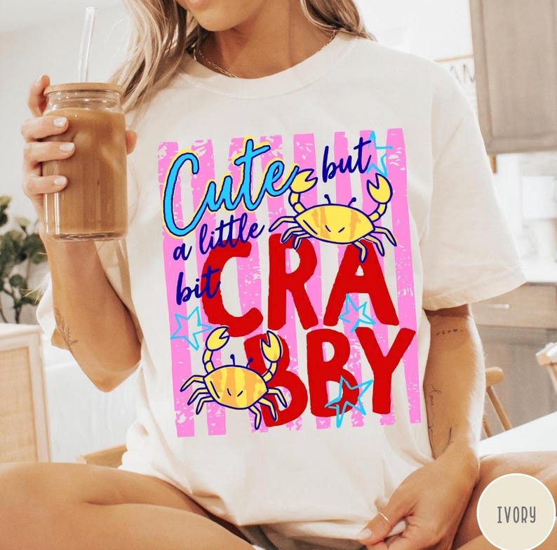 May include: An ivory t-shirt featuring a graphic print with pink vertical stripes, blue stars, and two yellow crabs. The text reads "Cute but a little bit CRABBY" in blue and red lettering. The design is centered on the front of the garment.