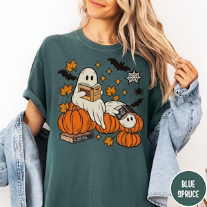 K&ouml;nnte beinhalten: Ein dunkelblaugr&uuml;nes T-Shirt mit einer Grafik von zwei wei&szlig;en Geistern, die B&uuml;cher lesen und auf orangefarbenen K&uuml;rbissen sitzen. Das Design enth&auml;lt orangefarbene Ahornbl&auml;tter, schwarze Flederm&auml;use, ein Spinnennetz und Sterne.