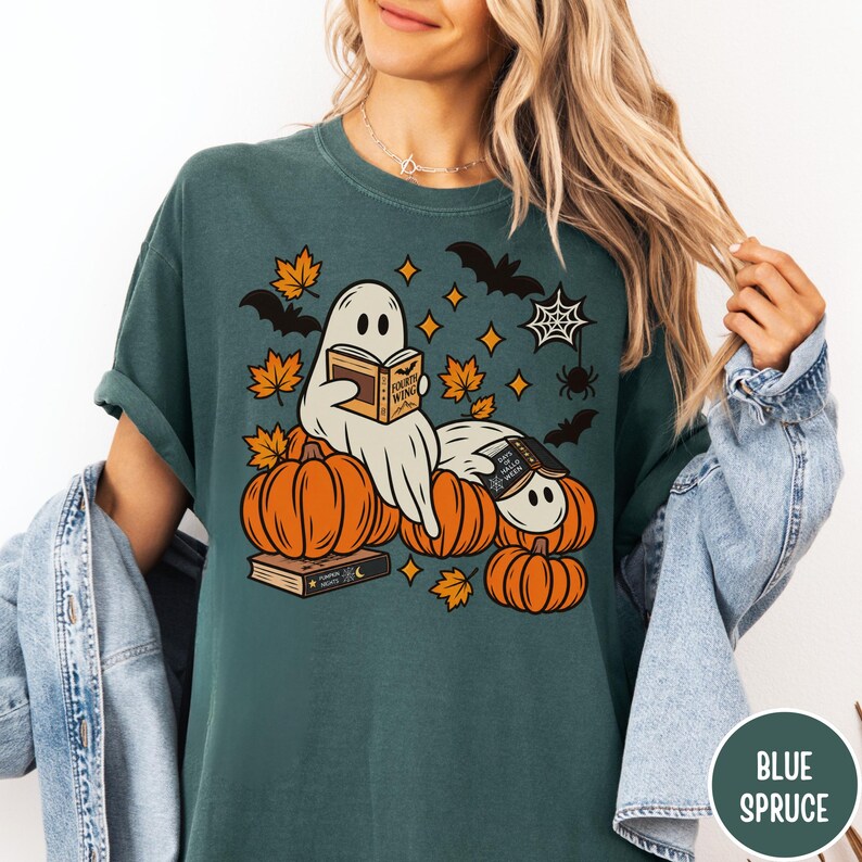 K&ouml;nnte beinhalten: Ein dunkelblaugr&uuml;nes T-Shirt mit einer Grafik von zwei wei&szlig;en Geistern, die B&uuml;cher lesen und auf orangefarbenen K&uuml;rbissen sitzen. Das Design enth&auml;lt orangefarbene Ahornbl&auml;tter, schwarze Flederm&auml;use, ein Spinnennetz und Sterne.